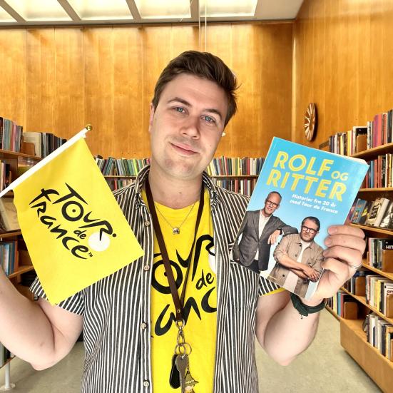 Bibliotekar Mathias Lyngs står med Tour de France t-shirt og et Tour de France-flag og holder Rolf og Ritters bog.